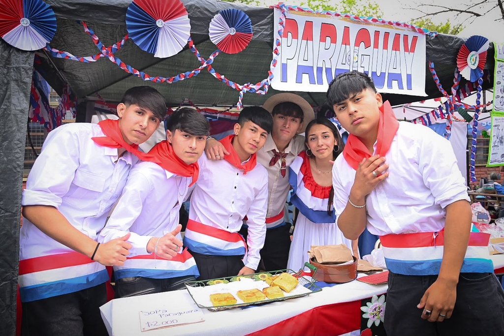 EMOCIONANTE | La Secundaria N° 2 celebró la 11° edición de la Feria de las Colectividades: Un viaje al corazón de nuestras raíces 4 Grupo de estudiantes posando con vestimenta típica y pañuelos rojos en el stand de Paraguay con platos regionales.