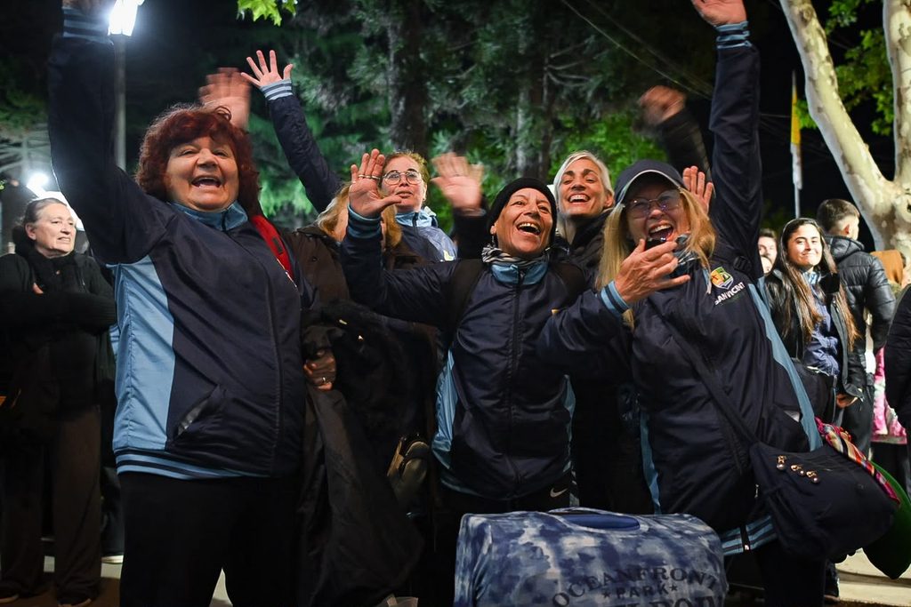 FINALES 2026 | San Vicente partió rumbo a las finales de los Juegos Bonaerenses 3 Participantes y adultos mayores de San Vicente saludando sonrientes desde la vereda con entusiasmo adentro del micro de viaje
